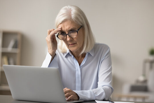 Troubled over business. Concerned worried senior lady accountant entrepreneur in glasses sit at office desk study report on notebook screen pondering company debt project bankruptcy financial problems