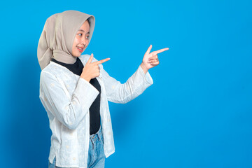 Confident young Asian Muslim woman in hijab smiling and pointing to the side with both hands. Happy facial expression suitable for promotional and advertising purposes.