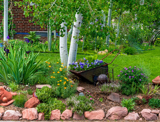 A mixed garden setting bordered by rocks with decorative elements including a wheelbarrow filled with pansies, a variety of flowers herbs and succulents and a Quacking Aspen Tree.