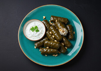 Stuffed grape leaves featuring rice, pine nuts, and herbs, plated on vibrant teal porcelain. Tzatziki sauce on the side, presented top-down over a solid black background in HD.