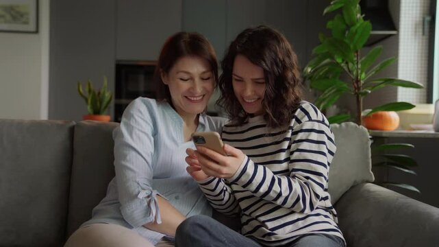 smiling mom and teenage daughter with acne bonding over funny content on smartphone. cozy moment on sofa bright kitchen, relaxed weekend vibes, real family connection at home. video call with family
