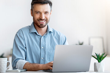 Smiling Young Businessman Sitting At Desk With Laptop In Modern Office, Looking At Camera, Free Space