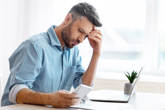 Worried male freelancer looking at smartphone screen while working at home office, received bad news, got scam message, thoughtful man touching forehead with concerned face expression, copy space