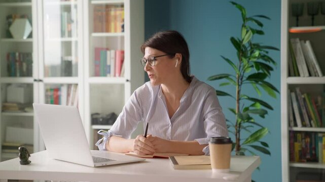 Beautiful mature woman working remotely on laptop, engaging in virtual communication with team, coach, manager. Professional video conference call at home or stylish office. Collaboration productivity