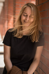 Fototapeta premium Portrait of young woman leaning against a brick wall, wearing black shirt and has long blonde hair that is blowing in the wind, the overall mood of the image is peaceful and serene.