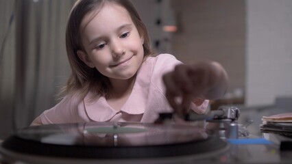Girl carefully places a needle on a vinyl record, exploring a vintage turntable with curiosity and...