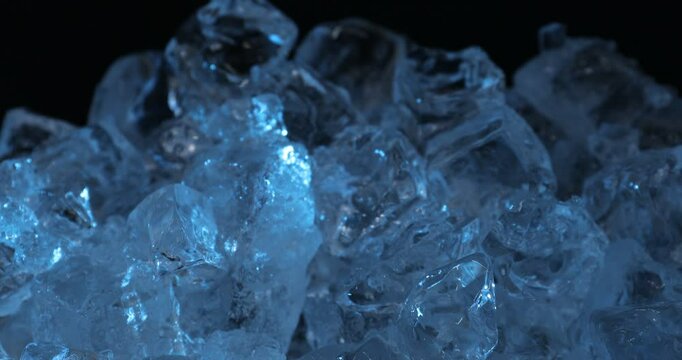 Many crystal clear ice cubes in blue light on black background, closeup. Camera moving up
