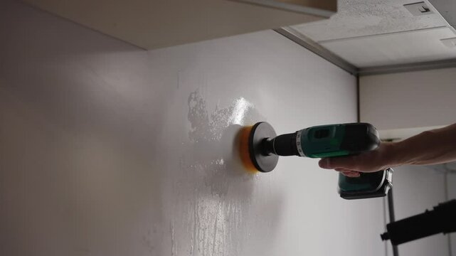 Cleaning service employee male wiping greasy kitchen wall surface using steam cleaner and rotating screwdriver brush for thorough surface sanitization and stain removal. Concept of housekeeping.