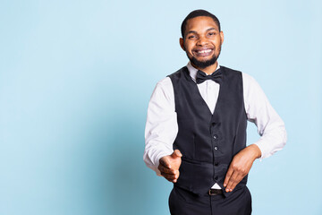 Bellboy hotel porter welcoming guests at the five star lodging resort, smiling and greeting people against blue background. Employee wearing a tuxedo and tie, tourism industry.