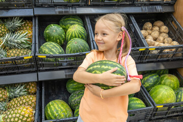 Fototapeta premium Cheerful little blonde girl holds a watermelon in fresh food supermarket. She stands in fruits and vegetables section with smile. Healthy eating. Family grocery shopping