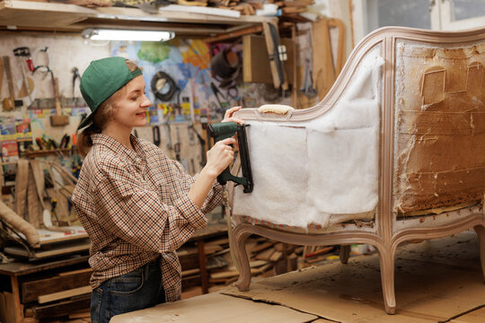 Female upholsterer using staple gun while restoring a vintage chair in a cozy workshop.