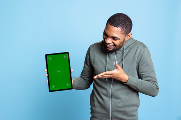 Tech content creator holding isolated green screen tablet doing promotion, talking with audience. Young adult pointing towards chroma key device, doing advertising against studio backdrop.