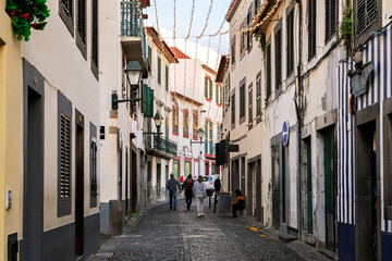 Obraz premium Pedestrians walk along the narrow cobblestone streets at the Rua de Santa Maria in the touristic center of the old town of Funchal, Madeira Island Portugal.
