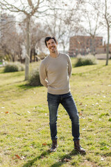 Full-body portrait of a cheerful man standing outdoors with hands behind his back, slightly tilted pose. Radiates friendliness, authenticity and joy. Vertical image, casual look, nature setting.