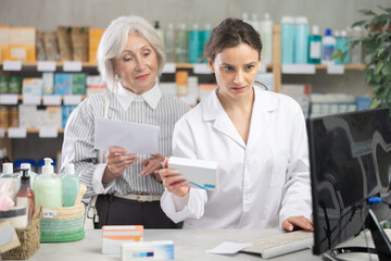 Obraz premium Female pharmacist holds a package of tablets and checks for other medications for an elderly woman at the pharmacy. Client consults with the pharmacist about the choice of pills in the pharmacy
