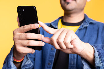 Adult Indian man with a man bun casually using his smartphone, browsing through online apps and social media. Guy with relaxed facial expression shows enjoyment against studio background. Close up.