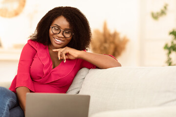 Online Chat. Smiling african woman in glasses using her laptop computer sitting on couch, copy...