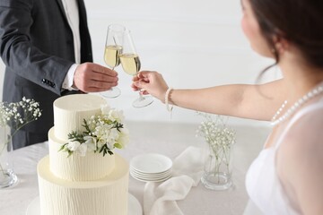Married couple clinking glasses of champagne at table with wedding cake indoors, closeup
