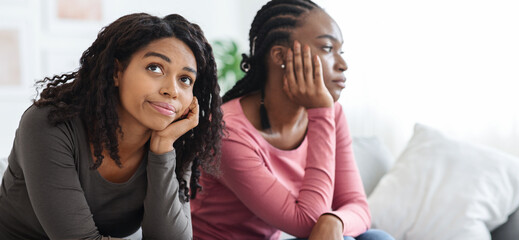Two sad african american women sitting apart on sofa at home, had quarrel, copy space. Frustrated...