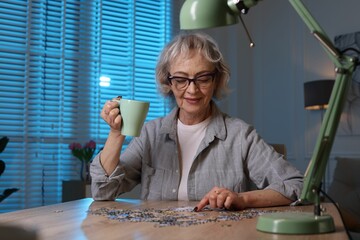 Woman with cup of drink solving jigsaw puzzle at wooden table indoors in evening