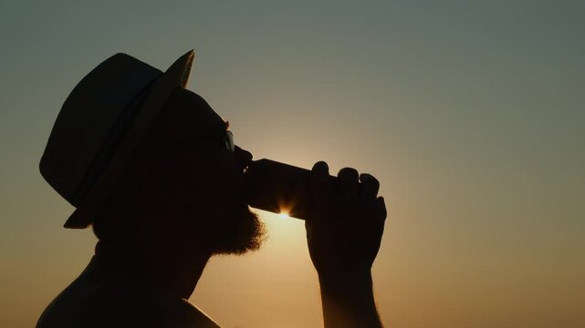 Adult bearded man in a hat drinking beer from a can by the sea at sunset. A refreshing sip of cold drink on a hot summer evening on vacation.