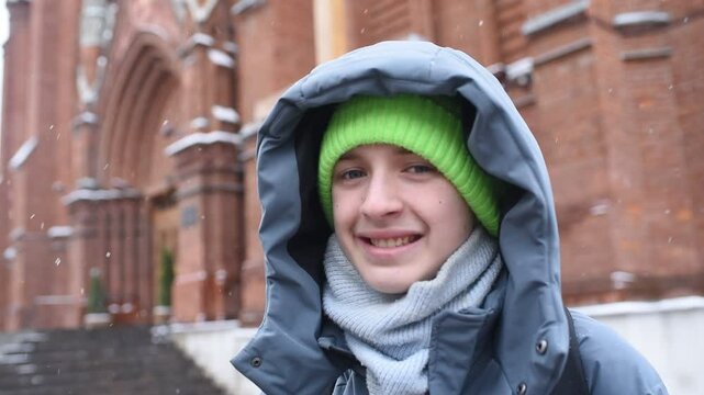 Portrait of smiling catholic boy in winter clothes in front of catholic church and cross on snowy day
