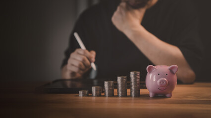 Young person plans finances with a tablet and pen while stacks of money coins and a pink piggy bank symbolize savings, investment, and financial growth on a wooden table.