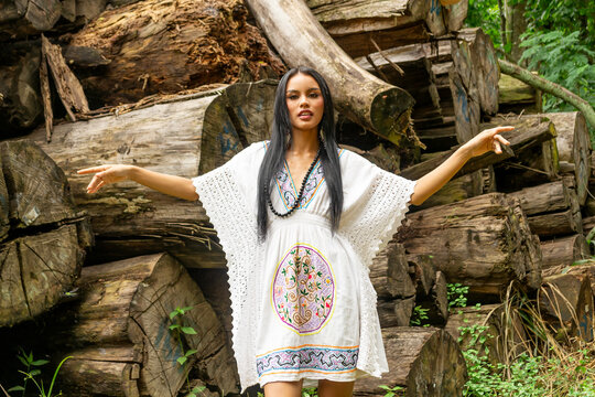 Amazon rainforest woman posing in a white dress with native Shipibo details, background of tree trunks felled by deforestation