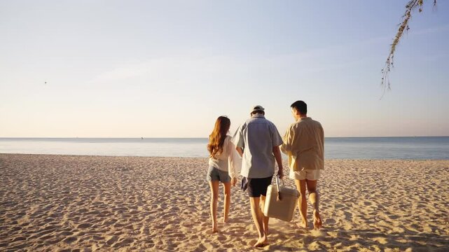 Happy man and woman travel the sea on summer beach holiday vacation. Group of Asian people friends enjoy and fun outdoor lifestyle holding cooler box walking to picnic on tropical beach in sunny day.