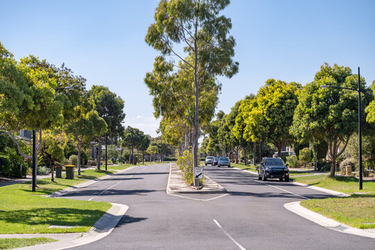 A suburban intersection with a straight asphalt road framed by mature trees and manicured lawns on the median and nature strip.A clean, well-defined street in a neighborhood.Williams Landing,Australia
