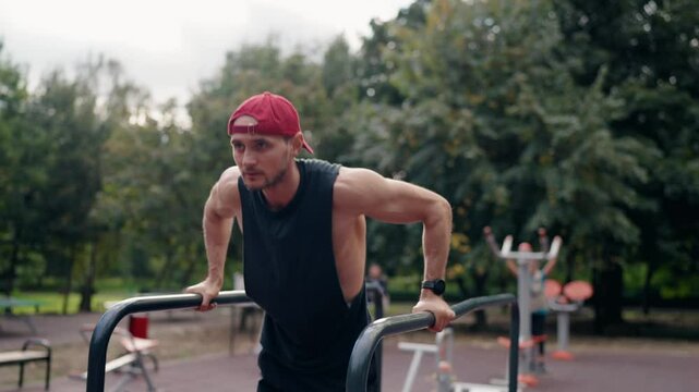 A young, fit man performs dips on parallel bars in an outdoor park setting. He wears a black tank top, shorts, and red baseball cap. Focused and determined, he strengthens his upper body.