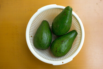 Three unripe avocado fruits inside a white basket on the wooden table.