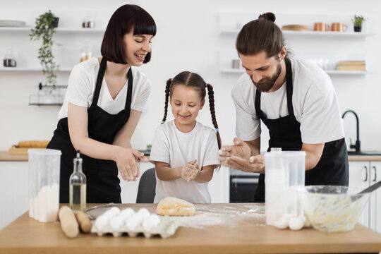 Smiling young parents and daughter in kitchen making dough, sharing happy bonding moment together