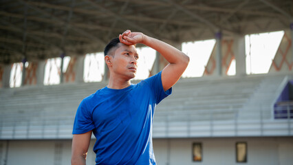 Asian man in blue t shirt is standing and wiping his sweat with a hand, taking a rest after running.
