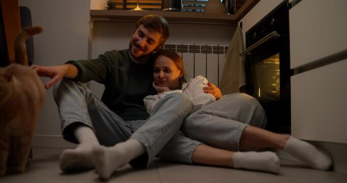 Happy blonde girl hugging her boyfriend lying on his lap while couple playing with cat sitting on floor at home romantic evening