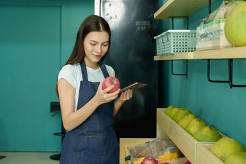 Young Caucasian woman holding red apple in hand while checking inventory with tablet inside fruit shop, managing small business confidently as owner preparing produce before opening store daily