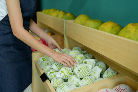 Closeup of female hands arranging green fruits wrapped in white protective netting on wooden shelf inside small fruit shop, representing hands-on owner care in running seasonal fresh produce business
