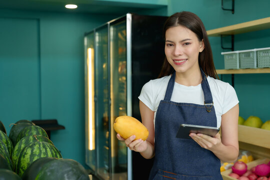 Caucasian woman holding ripe yellow mango and digital tablet while smiling confidently inside seasonal fruit shop, operating small business as owner with enthusiasm and fresh produce on display