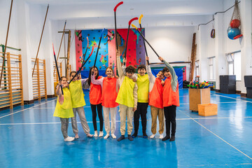Group of elementary school students wearing colorful vests holding hockey sticks up in the air,...