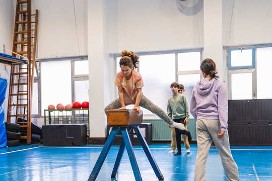 Young girl practicing gymnastics, jumping over a pommel horse in a school gym, watched by her classmates and instructor