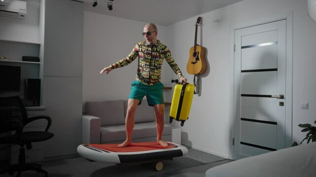 A middle-aged man in a colorful outfit, sunglasses, and a hat trains at home before his vacation. He balances on a board with a yellow suitcase in one hand and a smartphone in the other.