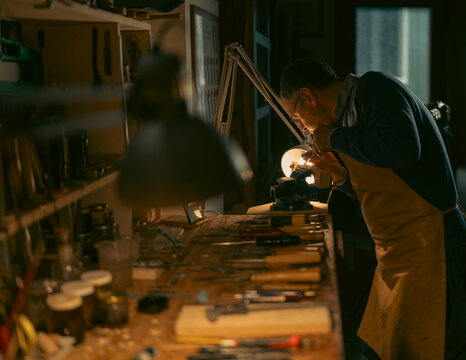 Skilled craftsman carefully carving wooden violin part, clamped in workbench vise, using specialized gouge under warm workshop lamp light