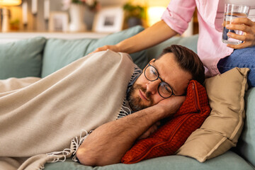 Sick man lying on the couch under a blanket, looking tired and unwell. He rests his head on a...