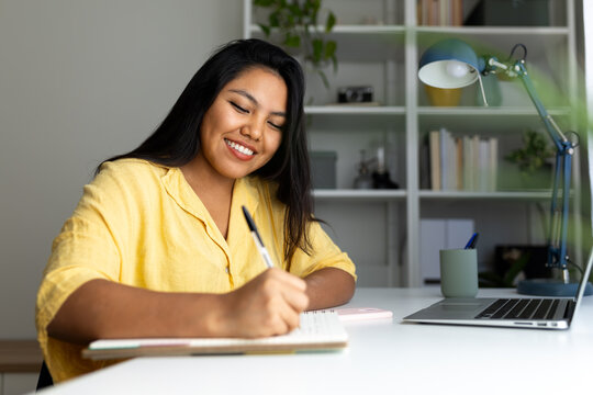 Latin american woman writing notes and smiling in home office