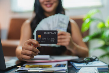Smiling businesswoman holding a credit card and cash while managing her finances at her home office desk, demonstrating financial security and online shopping