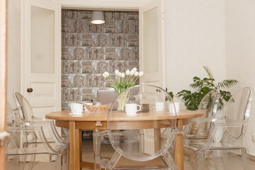 A cozy dining setup, featuring a wooden table with a laptop, tea set, and tulip centerpiece, surrounded by transparent chairs, exudes warmth and modern aesthetics.