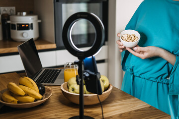 Woman holding a bowl of nuts in front of a ring light, laptop, and healthy food setup. Wellness...