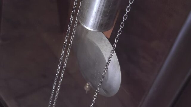Slowly swinging shiny metal pendulum of a large wall or floor clock with weight and chain for winding the gear mechanism. Closeup. Slow motion