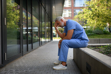 Fototapeta premium Frustrated and burnt-out doctor sitting outside the hospital, feeling down.