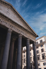 Obraz premium Low angle view of the pantheon's facade with its impressive roman columns and latin inscription, standing tall against a backdrop of a blue sky with wispy clouds and a modern building